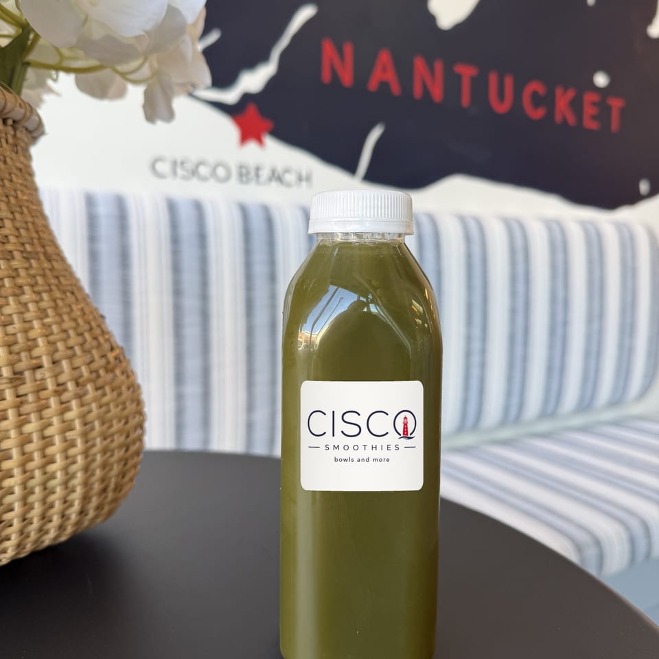 Cisco Smoothies Ginger Greens bottle on a table with striped seating and a Nantucket wall map behind it.