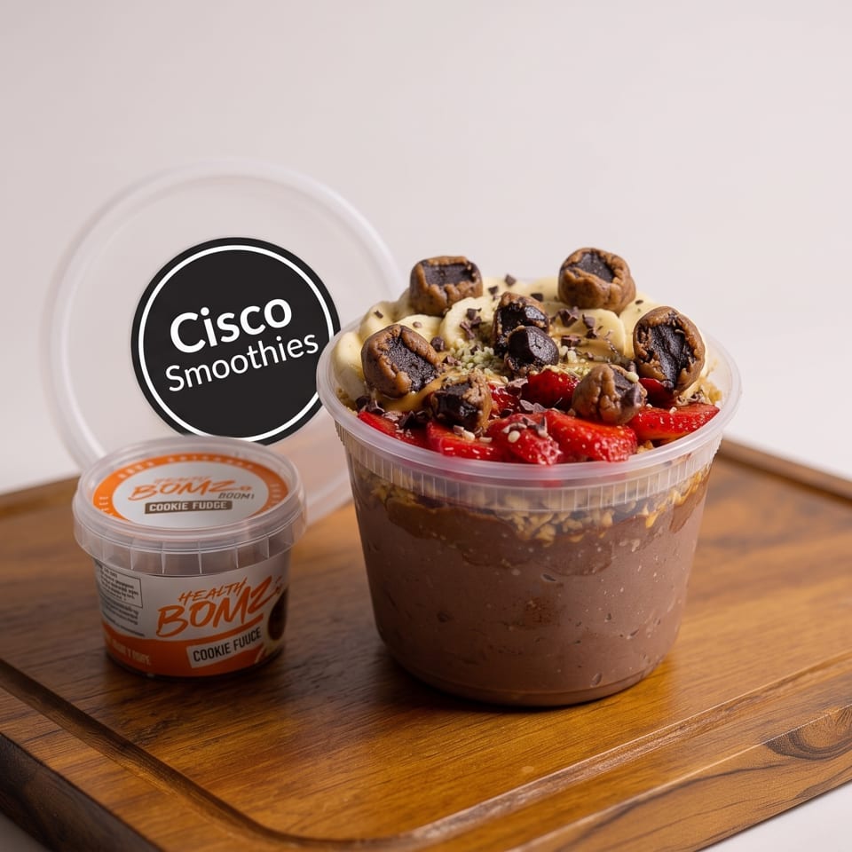 Chocolate smoothie bowl topped with bananas, strawberries, cacao nibs, and cookie pieces beside a cookie fudge container on a wooden board.
